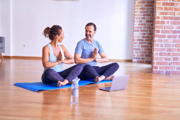 Middle age beautiful sporty couple smiling happy. Sitting on mat practicing yoga doing prayer pose using laptop at gym - Photo, Image