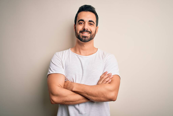 Young handsome man with beard wearing casual t-shirt standing over white background happy face smiling with crossed arms looking at the camera. Positive person. - Photo, Image