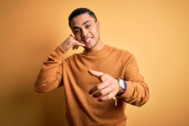 Young brazilian man wearing casual sweater standing over isolated yellow background smiling doing talking on the telephone gesture and pointing to you. Call me. - Photo, Image