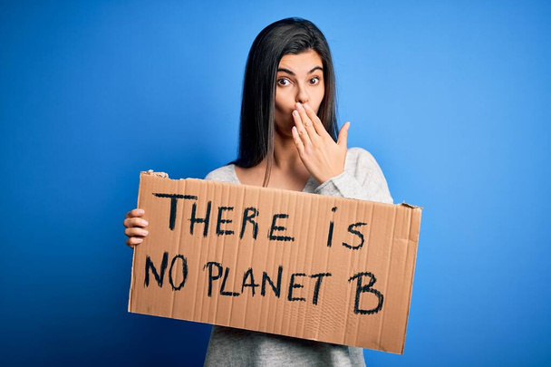 Jeune belle militante brune tenant une bannière protestant pour prendre soin de la planète couvrir la bouche avec la main choquée par la honte pour erreur, expression de la peur, effrayée dans le silence, concept secret
 - Photo, image