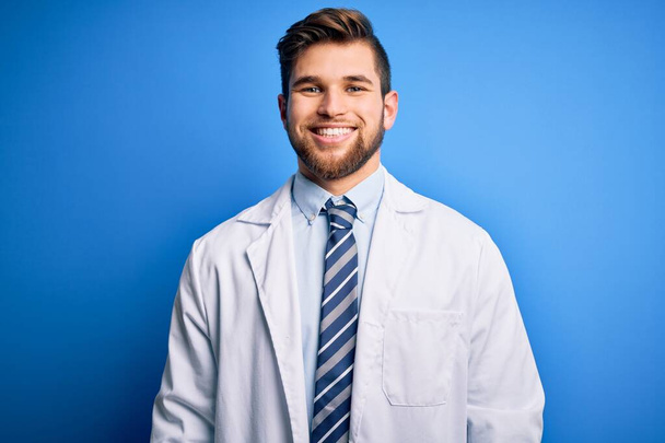 Young blond therapist man with beard and blue eyes wearing coat and tie over background with a happy and cool smile on face. Lucky person. - Fotó, kép