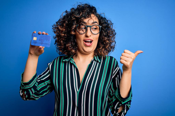 Jeune belle femme arabe bouclée portant des lunettes tenant l'argent de carte de crédit pour le pointage de paiement et montrant avec le pouce sur le côté avec sourire heureux visage
 - Photo, image