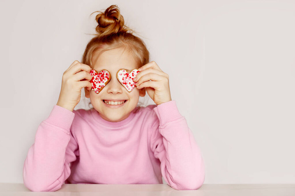 Een meisje in een roze trui houdt peperkoek vast in de vorm van een hart. Het kind sluit zijn ogen met koekjes - Foto, afbeelding
