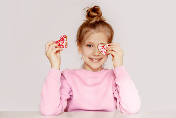 Uma menina de camisola rosa está segurando pão de gengibre na forma de um coração. A criança fecha os olhos com biscoitos
 - Foto, Imagem
