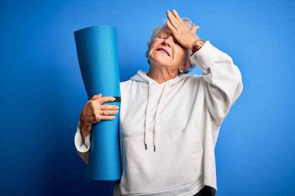 Senior hermosa mujer deportiva que sostiene la estera para el yoga de pie sobre fondo azul aislado estresado con la mano en la cabeza, sorprendido con la vergüenza y la cara sorpresa, enojado y frustrado. Miedo y malestar por error
. - Foto, Imagen