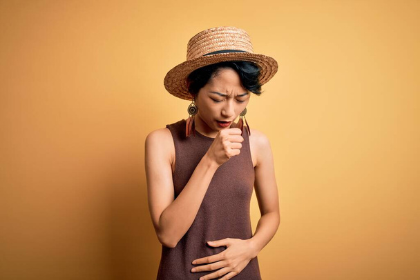 Jeune belle fille asiatique portant un t-shirt décontracté et un chapeau sur fond jaune isolé se sentant mal à l'aise et toussant comme symptôme de rhume ou de bronchite. Concept de soins de santé
. - Photo, image