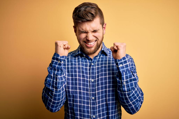 Young blond businessman with beard and blue eyes wearing shirt over yellow background excited for success with arms raised and eyes closed celebrating victory smiling. Winner concept. - Fotoğraf, Görsel