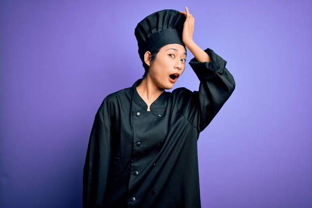 Joven hermosa mujer chef chino con uniforme de cocina y sombrero sobre fondo púrpura sorprendido con la mano en la cabeza por error, recordar el error. Olvidado, mal concepto de memoria
. - Foto, imagen