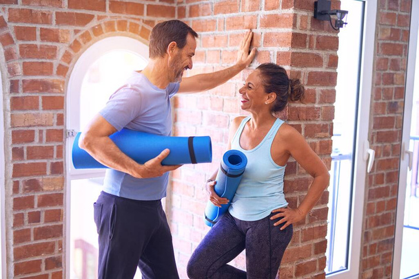 Middle age beautiful sporty couple smiling happy. Standing with smile on face holding mat and speaking at gym - Photo, Image