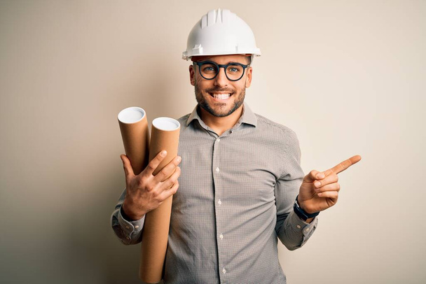 Junger Architekt mit Bauarbeiterhelm, der einen Plan aus Projektpapier vor isoliertem Hintergrund hält und sehr glücklich mit der Hand und dem Finger zur Seite zeigt - Foto, Bild