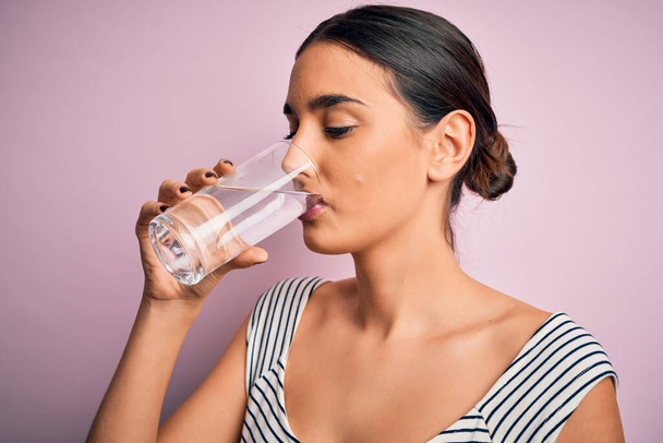 Young beautiful brunette woman drinking glass of healthy water to refreshment standing over isolated pink background - Foto, afbeelding