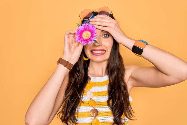Jonge mooie hippie vrouw met blauwe ogen dragen zonnebril houden roze bloem over oog gestrest met de hand op het hoofd, geschokt met schaamte en verrassing gezicht, boos en gefrustreerd. Angst en overstuur voor fouten. - Foto, afbeelding