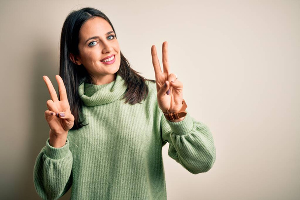 青い目をした若いブルネットの女性は、勝利のサインをして指を示すカメラを見て笑って白い背景にタートルネックセーターを着ていた。第二番. - 写真・画像