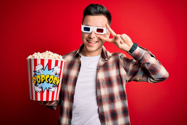Mladý pohledný běloch nosí 3D brýle a jí popcorn, dělá mírový symbol s prsty přes obličej, vesele se usmívá, ukazuje vítězství - Fotografie, Obrázek