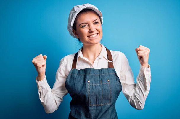 Young beautiful baker woman with blue eyes wearing apron and cap over blue background very happy and excited doing winner gesture with arms raised, smiling and screaming for success. Celebration concept. - Photo, Image