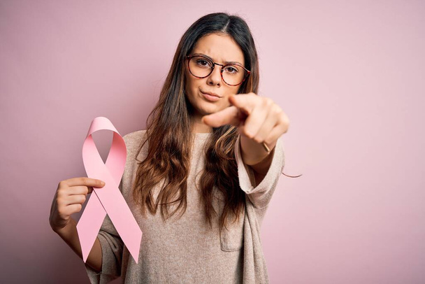 Mladá krásná bruneta žena v brýlích drží růžovou stužku proti rakovině ukazující prstem na fotoaparát a na vás, znak ruky, pozitivní a sebevědomé gesto zepředu - Fotografie, Obrázek