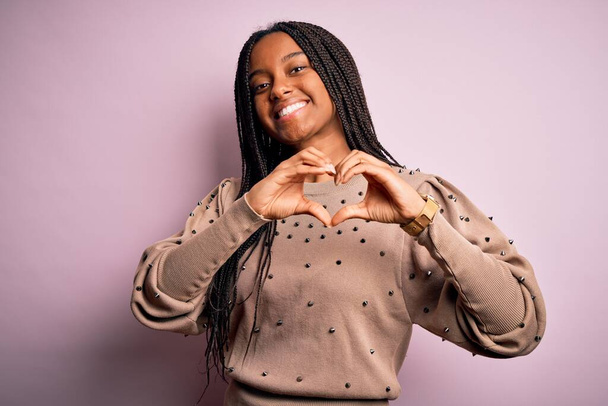Mujer afroamericana joven con suéter de moda sobre fondo rosa aislado sonriendo en el amor mostrando el símbolo del corazón y la forma con las manos. Concepto romántico
. - Foto, Imagen