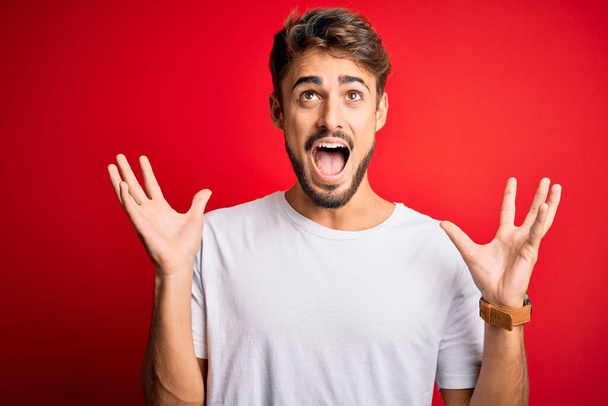 Young handsome man with beard wearing casual t-shirt standing over red background celebrating mad and crazy for success with arms raised and closed eyes screaming excited. Winner concept - Fotoğraf, Görsel