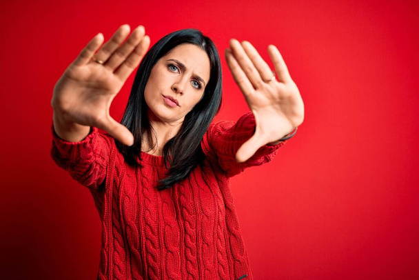 Mladá brunetka žena s modrýma očima na sobě ležérní svetr na izolovaném červeném pozadí dělat rám pomocí dlaně a prsty, kamera perspektiva - Fotografie, Obrázek