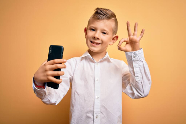 Fiatal kis kaukázusi gyerek használ okostelefon és nézi a képernyőn a telefon csinál oké jel ujjakkal, kiváló szimbólum - Fotó, kép