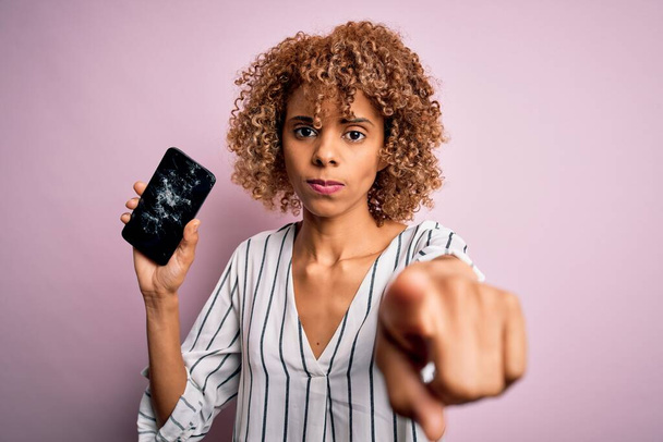 Junge afrikanisch-amerikanische lockige Frau hält kaputtes Smartphone und zeigt mit dem Finger auf die Kamera und auf dich, Handzeichen, positive und selbstbewusste Geste von vorne - Foto, Bild