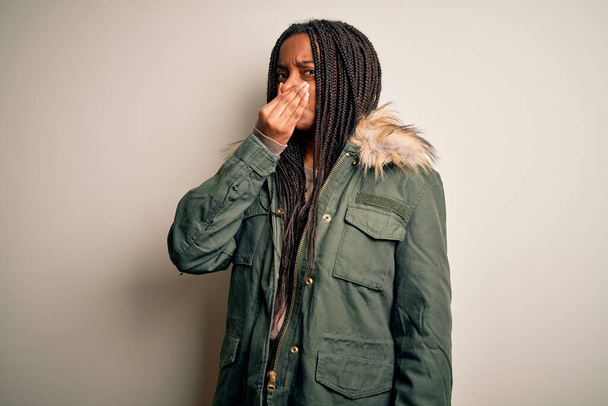 Young african american woman wearing winter parka coat over isolated background smelling something stinky and disgusting, intolerable smell, holding breath with fingers on nose. Bad smell - Fotoğraf, Görsel