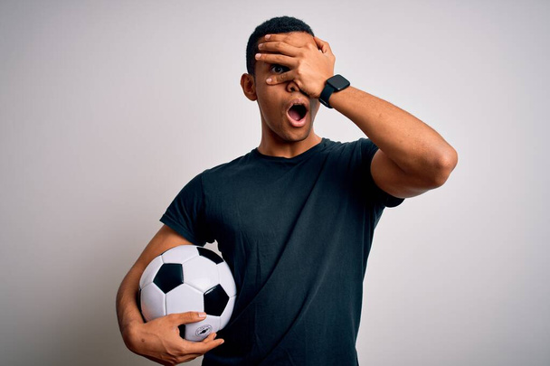 Yakışıklı Afro-Amerikan adam futbol topunu beyaz arka planda tutarak oynuyor. Şok olmuş bir şekilde yüzünü ve gözlerini kapatıyor. Utanmış bir ifadeyle parmaklarından bakıyor.. - Fotoğraf, Görsel