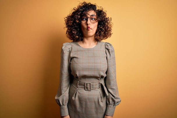 Beautiful arab business woman wearing dress and glasses standing over yellow background making fish face with lips, crazy and comical gesture. Funny expression. - Valokuva, kuva