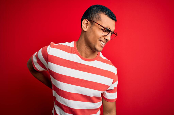 Fiatal, jóképű afro-amerikai férfi alkalmi csíkos pólót és szemüveget visel Hátfájás, kézfogdosás, izomfájdalom - Fotó, kép