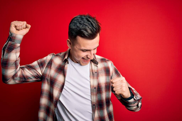 Young handsome caucasian man wearing casual modern shirt over red isolated background Dancing happy and cheerful, smiling moving casual and confident listening to music - Фото, изображение