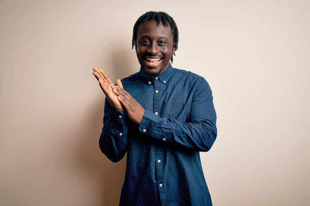 Νεαρός όμορφος Αφροαμερικάνος που φοράει casual πουκάμισο στέκεται πάνω από λευκό φόντο χειροκροτώντας χαρούμενα και χαρούμενα, χαμογελαστά περήφανα χέρια μαζί - Φωτογραφία, εικόνα