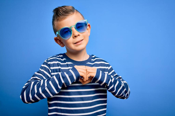 青い目の若い白人の子供は、ハートのシンボルと手で形を示す愛に笑みを浮かべて青い背景にサングラスをかけて立っています。ロマン主義. - 写真・画像