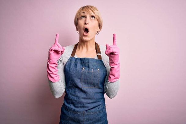 Jonge blonde schonere vrouw met kort haar dragen schort en handschoenen over roze achtergrond verbaasd en verrast omhoog kijken en wijzen met vingers en opgeheven armen. - Foto, afbeelding