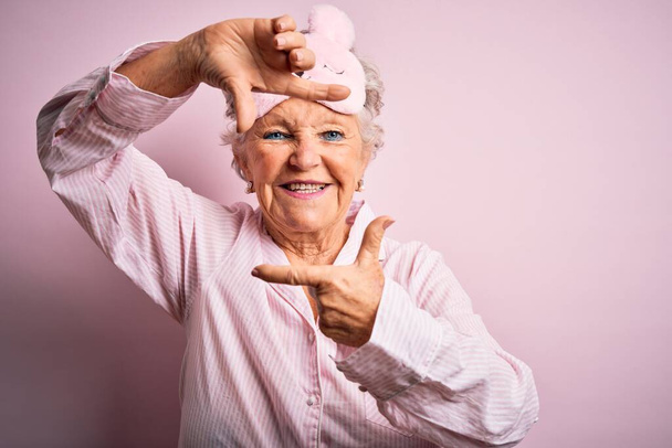 Senior beautiful woman wearing sleep mask and pajama over isolated pink background smiling making frame with hands and fingers with happy face. Creativity and photography concept. - Photo, Image