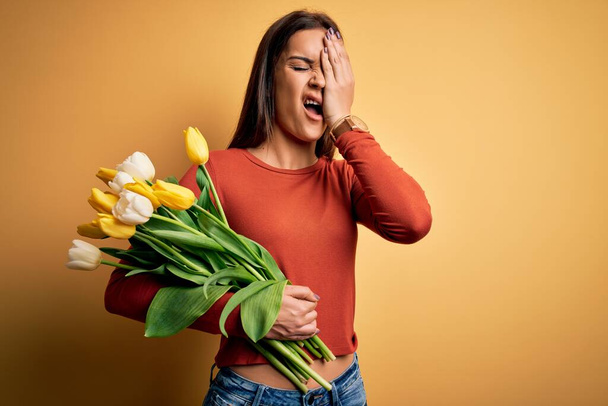 Jeune belle femme brune tenant un bouquet de fleurs de tulipes sur fond jaune Bâillement fatigué couvrant la moitié du visage, des yeux et de la bouche avec la main. Le visage souffre dans la douleur
. - Photo, image