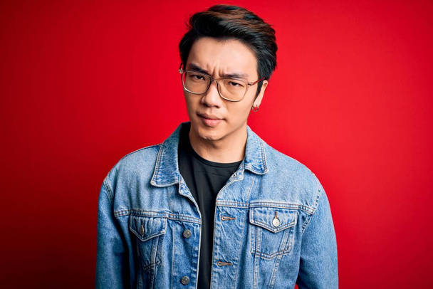 Junger gutaussehender chinesischer Mann in Jeansjacke und Brille über rotem Hintergrund skeptisch und nervös, die Stirn runzelnd wegen des Problems. Negative Person. - Foto, Bild