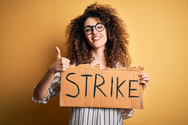 Junge schöne Aktivistin mit lockigem Haar und Piercing protestiert im Streik mit Plakat glücklich mit breitem Lächeln tun ok Zeichen, Daumen nach oben mit den Fingern, hervorragendes Zeichen - Foto, Bild