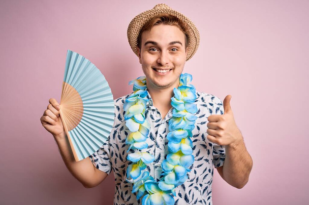 Joven guapo pelirrojo turista de vacaciones vistiendo lei hawaiano y sombrero usando ventilador de mano sonriendo feliz y positivo, pulgar hacia arriba haciendo excelente y signo de aprobación
 - Foto, imagen