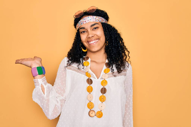 Young african american curly hippie woman wearing sunglasses and vintage accessories smiling cheerful presenting and pointing with palm of hand looking at the camera. - Фото, зображення
