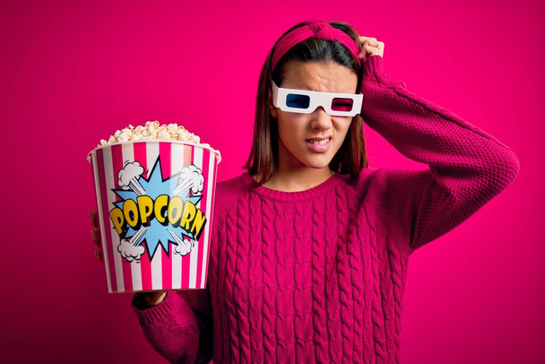 Young beautiful girl watching movie using 3d glasses eating box with popcorns confuse and wondering about question. Uncertain with doubt, thinking with hand on head. Pensive concept. - Photo, Image