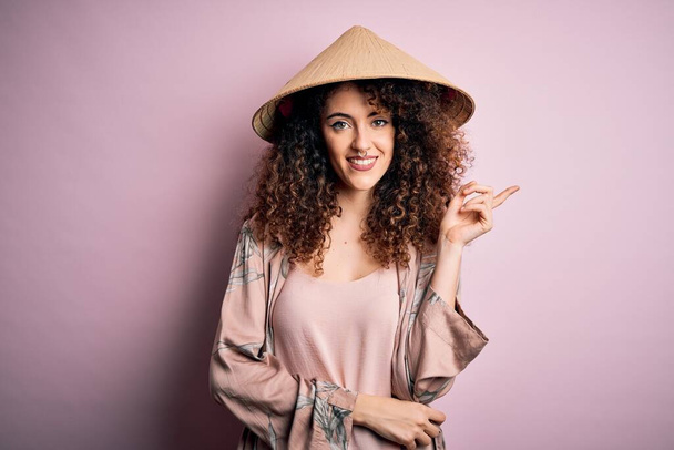 Fiatal gyönyörű nő göndör hajjal és piercing visel hagyományos ázsiai kúpos kalap nagy mosollyal az arcán, mutató kézzel és ujjal oldalra néz a kamerába. - Fotó, kép