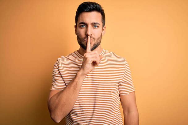 Young handsome man with beard wearing casual striped t-shirt over yellow background asking to be quiet with finger on lips. Silence and secret concept. - Photo, Image