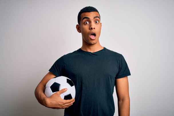 Przystojny Afroamerykanin gra w piłkę nożną trzymając piłkę na białym tle w szoku twarzy, sceptyczny i sarkastyczny, zaskoczony otwartymi ustami - Zdjęcie, obraz