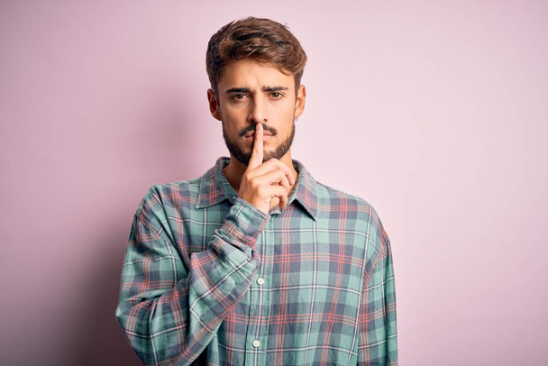 Young handsome man with beard wearing casual shirt standing over pink background asking to be quiet with finger on lips. Silence and secret concept. - Photo, Image