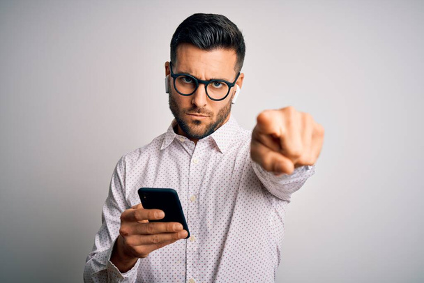 Young handsome man wearing glasses having conversation using smartphone and earphones pointing with finger to the camera and to you, hand sign, positive and confident gesture from the front - Photo, Image