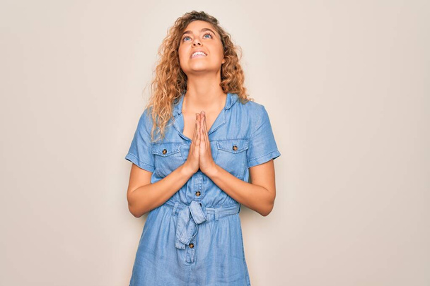 Young beautiful woman with blue eyes wearing casual denim dress over white background begging and praying with hands together with hope expression on face very emotional and worried. Begging. - Photo, Image