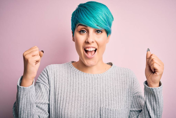 Young beautiful woman with blue fashion hair wearing casual sweater over pink background celebrating surprised and amazed for success with arms raised and open eyes. Winner concept. - Foto, Bild