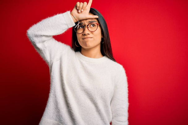 Mladý krásný asijské žena na sobě ležérní svetr a brýle přes červené pozadí dělat legraci z lidí s prsty na čele dělá poražený gesto posměch a urážlivé. - Fotografie, Obrázek