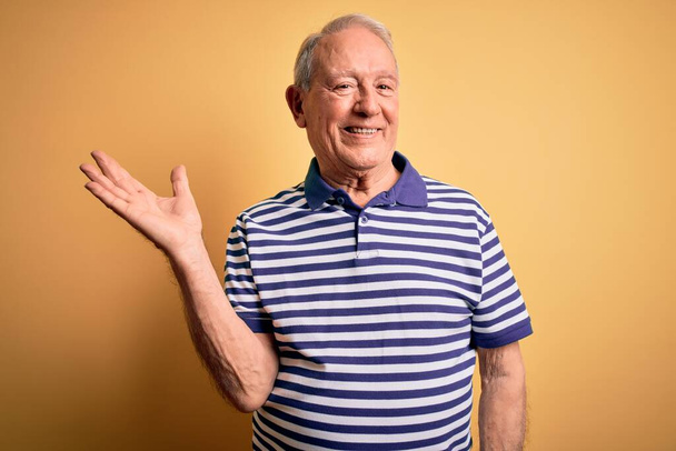 Hombre mayor de pelo gris que usa una camiseta casual de rayas navales de pie sobre fondo amarillo sonriendo alegre presentando y señalando con la palma de la mano mirando a la cámara
. - Foto, imagen