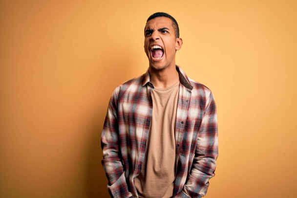 Young handsome african american man wearing casual shirt standing over yellow background angry and mad screaming frustrated and furious, shouting with anger. Rage and aggressive concept. - Foto, Imagem